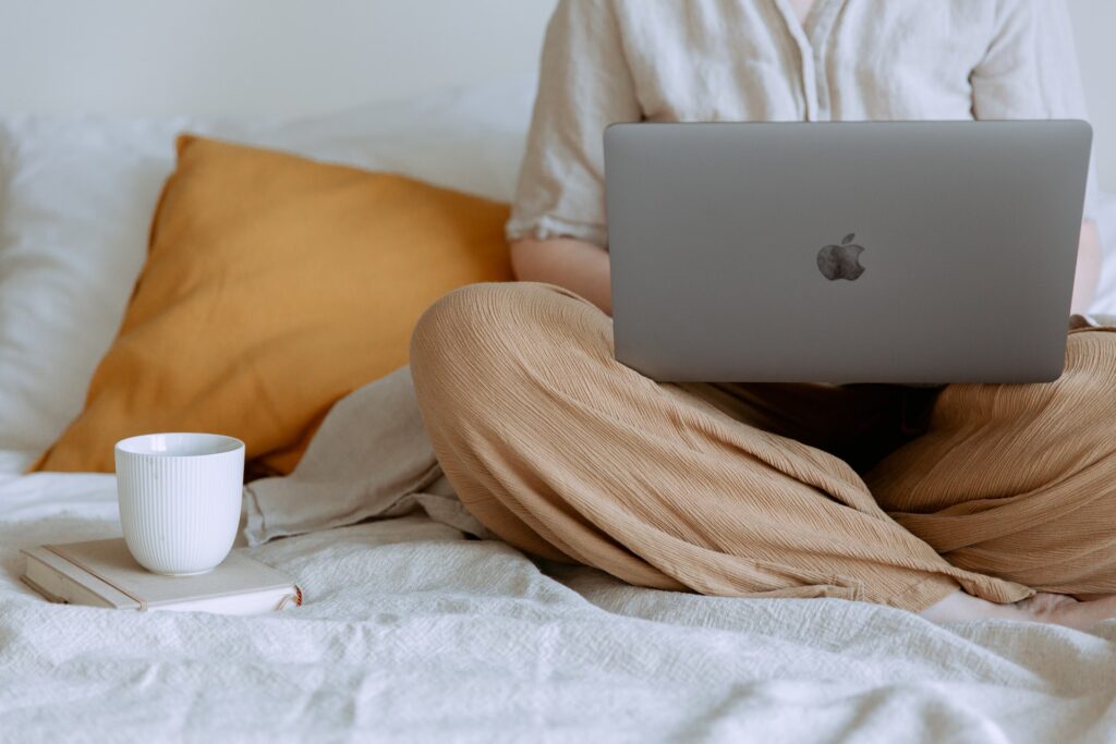 person sitting cross-legged on a bed, mac in lap, with a coffee cup and orange cushion beside